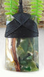 A resin pendant featuring a black braided wrap closure and a clear resin body filled with natural elements including green leaves, brown twigs, and white flower petals, shown against a background of faux ferns and wood.