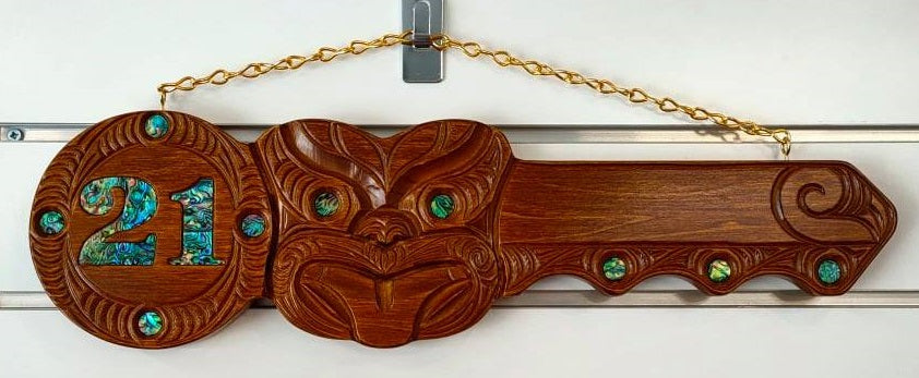 A wooden 21st key carving with Paua inlay, hanging against a white background.